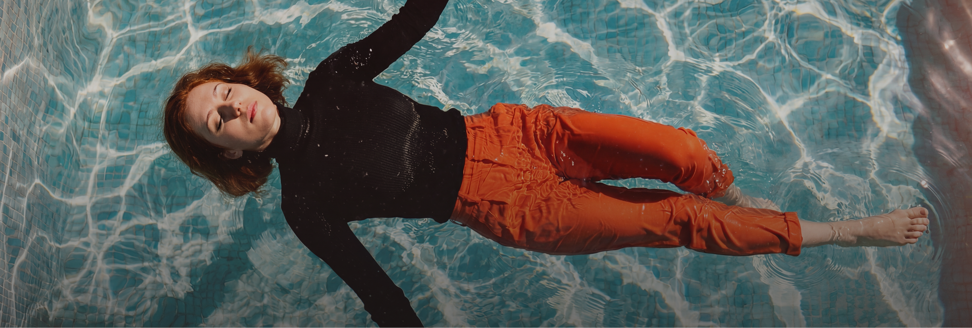 Draufsicht auf eine rothaarige Frau, die entspannt und mit geschlossenen Augen im klaren, hellblauen Wasser eines Pools schwebt. Sie trägt einen schwarzen Langarm-Rollkragenpullover und eine leuchtend orangefarbene Hose. Das Sonnenlicht erzeugt tanzende Reflexionen auf der Wasseroberfläche und den hellen Mosaikfliesen am Boden des Beckens.