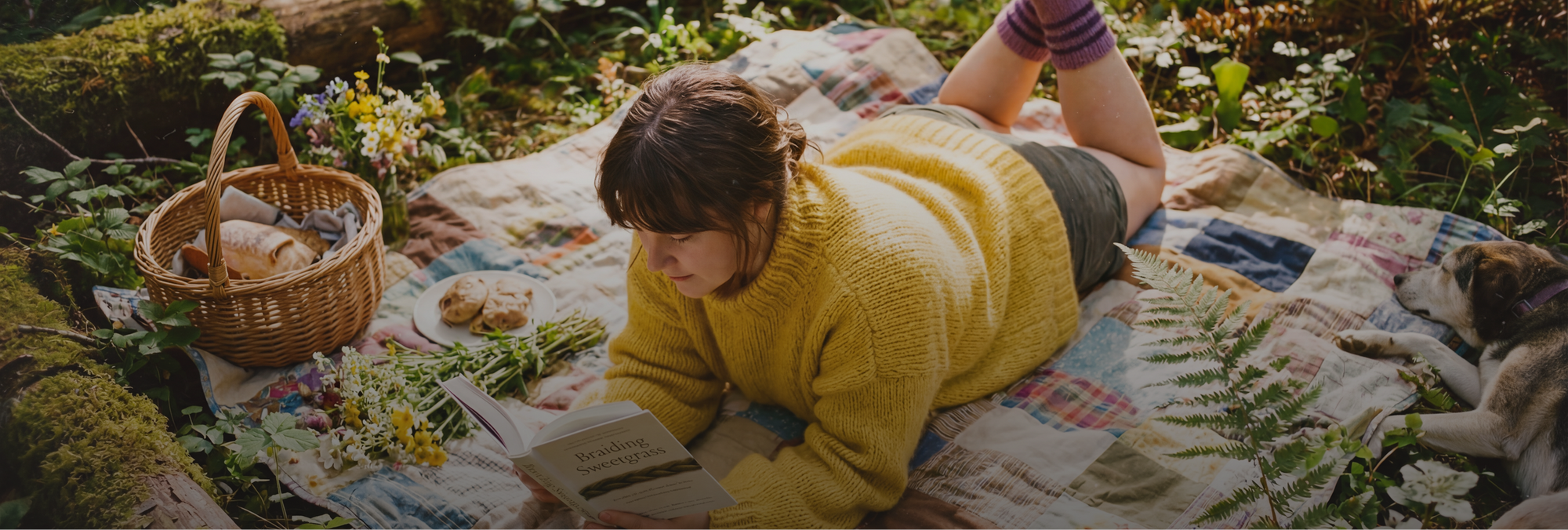 Eine junge Frau liegt auf dem Bauch auf einer bunten Patchwork-Decke im sonnendurchfluteten Wald und liest konzentriert das Buch „Braiding Sweetgrass“. Sie trägt einen grob gestrickten gelben Pullover und gestreifte lila Socken. Neben ihr stehen ein Picknickkorb mit Brot, ein Teller mit Gebäck und frische Wildblumen. Rechts im Bild schläft ein kleiner, braun-weißer Hund im weichen Schatten.